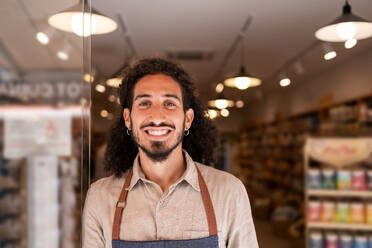 Portrait of positive bearded man in apron smiling and looking at camera while standing near dark blurred lighted background in supermarket - ADSF46328