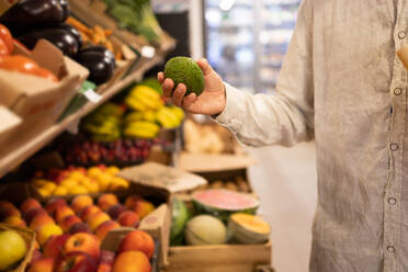 Crop anonymous person in casual clothes standing while choosing fruit by fruit shelves in grocery store with various fruits - ADSF46332