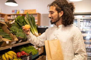 Portrait of young ethnic man with curly hair in casual clothes standing near shelves with assorted fresh vegetables and fruits while shopping indoors - ADSF46333