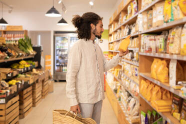 Positive side view young curly hair male customer in casual clothes standing near shelves with goods while shopping with bag in grocery shop - ADSF46335