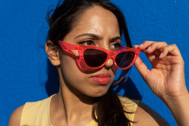 Portrait of serious young Indian female holding sunglasses on nose and looking away while standing in light with shadow in blue background wall - ADSF46336