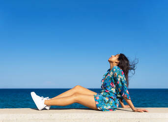 Side view of smiling young Indian female with long dark hair eyes closed relaxing on concrete surface while leaning on hands under cloudless blue sky - ADSF46337