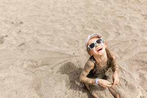 From above of smiling child in sunglasses and cap looking up while sitting with wet sand on shabby swimwear on beach in sunlight and enjoying summer vacation - ADSF46340