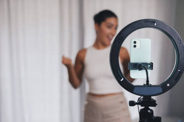 Soft focus of professional female influencer shooting video via cellphone on tripod with ring lamp at home - ADSF46341