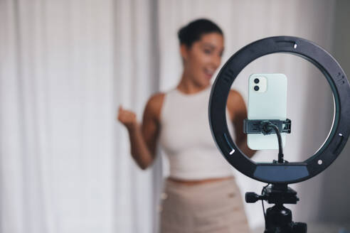 Soft focus of professional female influencer shooting video via cellphone on tripod with ring lamp at home - ADSF46341