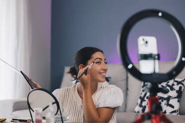 Young positive Hispanic female blogger applying eyeshadow on eyelid with brush while recording makeup tutorial for social media via smartphone - ADSF46343