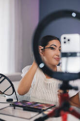 Young Hispanic female influencer shooting video on smartphone with ring lamp while applying mascara on eyelashes at home during beauty routine - ADSF46346