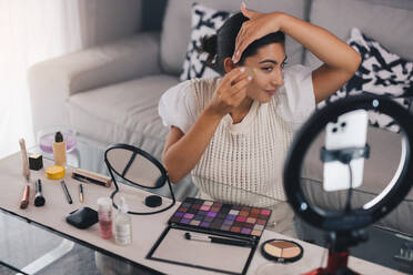 High angle of young Hispanic female influencer applying foundation on cheek with sponge while sitting at coffee table and recording video on cellphone - ADSF46347