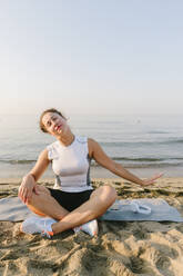 Woman stretching arm sitting on exercise mat in front of sea - SIF00776