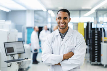 Confident technician smiling with arms crossed while colleagues are talking in electronics factory - DIGF20233