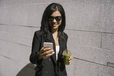Lächelnde Geschäftsfrau, die ein Smartphone benutzt und einen grünen Smoothie an der Wand hält - YBF00080