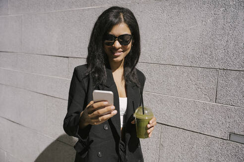 Smiling businesswoman using smart phone and holding green smoothie near wall - YBF00080