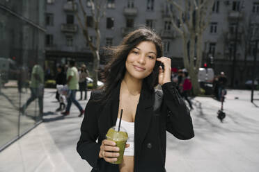 Lächelnde Geschäftsfrau mit grünem Smoothie in der Hand in der Stadt - YBF00083