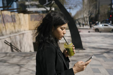 Businesswoman using smart phone and drinking green smoothie - YBF00084