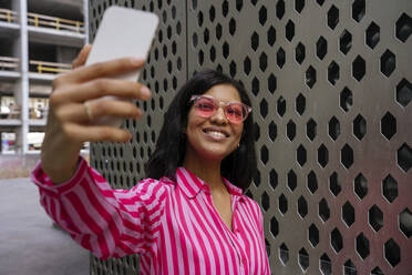 Happy woman wearing sunglasses and taking selfie through smart phone near wall - YBF00091