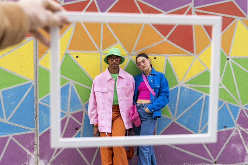 Cropped hand holding picture frame with friends standing in front of colorful wall - AFVF09318