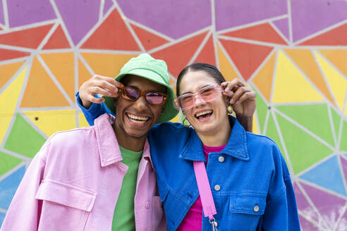 Cheerful couple holding sunglasses in front of colorful wall - AFVF09326