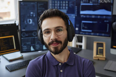 Lächelnder IT-Fachmann mit drahtlosen Kopfhörern vor einem Computer - KPEF00137