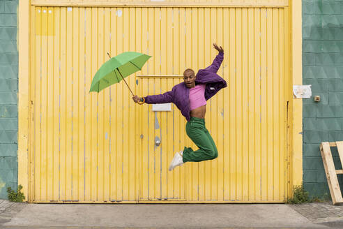 Non-binary person jumping with umbrella in front of yellow shutter door - AFVF09353