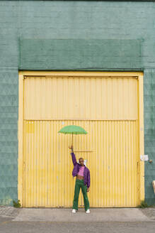 Nicht-binäre Person stehend mit Regenschirm vor einer gelben Rollladentür - AFVF09354