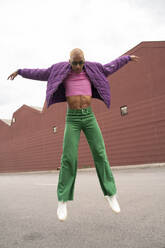 Non-binary person wearing sunglasses jumping in front of building - AFVF09375