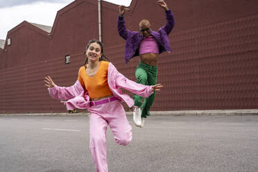 Cheerful woman dancing with friend jumping in front of building - AFVF09395