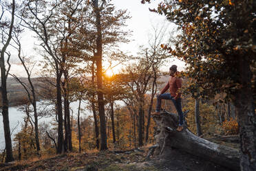 Contemplative man standing on tree trunk near lake - JOSEF20454