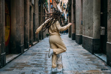 A woman with braids wearing a beige outfit and sneakers dances on a narrow street in the charming old town - ADSF46357