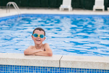 Ein fröhliches Kind mit Schwimmbrille steht am Pool, die Arme verschränkt, und genießt die Sommerpause im erfrischenden blauen Wasser - ADSF46359