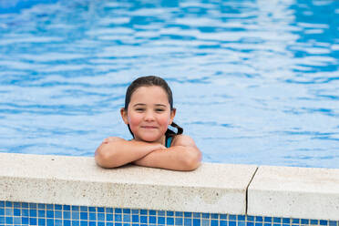 Ein fröhliches Kind mit feuchtem Haar steht am Pool und blickt mit verschränkten Armen in die Linse, während es den Sommerurlaub genießt - ADSF46360