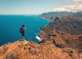 Ein waghalsiger Wanderer fängt die atemberaubende Aussicht auf die Berge und das azurblaue Meer Teneriffas von einer Klippe aus mit seiner Kamera ein - ADSF46375