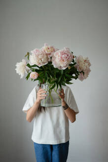 A bashful young girl hides behind colorful peonies, adding a pop of color to a simple background - ADSF46380