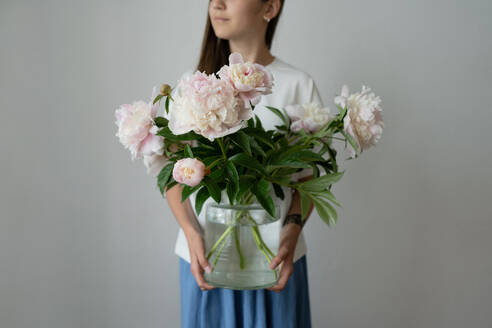 A young girl admires a stunning display of peonies in a glass jar against a neutral backdrop - ADSF46381