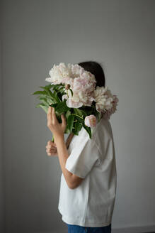 A young child playfully hides behind a beautiful display of blooming peonies set against a neutral gray wall - ADSF46382
