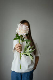 A young child playfully hides behind colorful peonies, dressed casually and enjoying a sunny day against a gray wall - ADSF46383