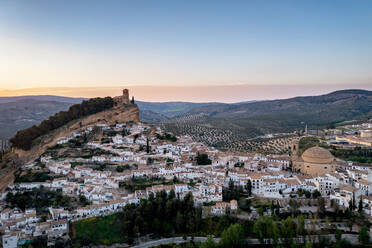 Malerischer Blick auf eine alte Stadt auf einem Hügel mit steinernen Gebäuden und grünen Bäumen unter wolkenlosem Himmel bei Sonnenuntergang im Dorf n Montefrio, Pueblos Blancos in Spanien - ADSF46384