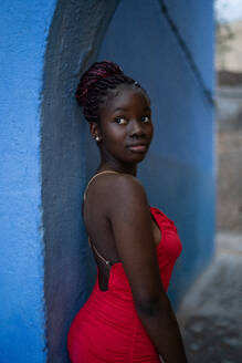 Eine nachdenkliche afroamerikanische Frau in einem auffälligen roten Kleid posiert vor einem ruhigen blauen Hintergrund und ist in Gedanken versunken - ADSF46392