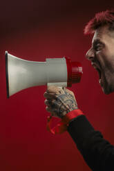 A fiery-haired man with tattooed hands passionately uses a megaphone in front of a bold red studio backdrop - ADSF46393