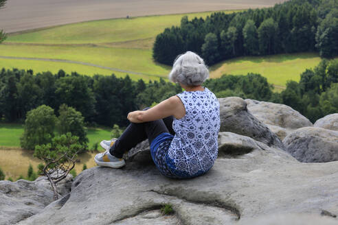Thoughtful mature woman sitting on rock in front of trees - JTF02363