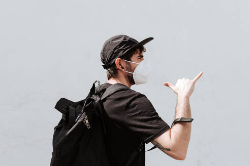 Amidst the pandemic, a trendy man dons a face mask and backpack while flashing a shaka sign - ADSF46441