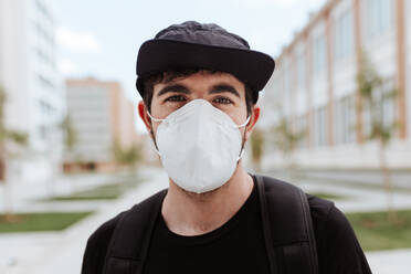 A fashionable man dons a face mask and cap while posing in front of buildings during the COVID-19 crisis. - ADSF46442