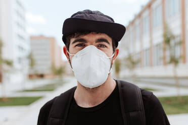 Ein modebewusster Mann setzt sich eine Gesichtsmaske und eine Mütze auf, während er während der COVID-19-Krise vor einem Gebäude posiert. - ADSF46442