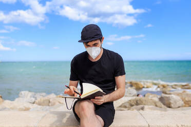 A man wearing a mask and cap sits on a fence, writing in his diary while enjoying the view of the ocean - ADSF46444
