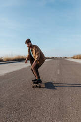 Ein eleganter bärtiger Skater gleitet auf seinem Skateboard eine ländliche Asphaltstraße hinunter - ADSF46447