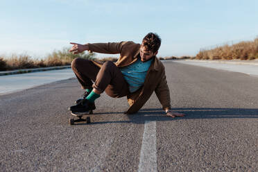 Ein modischer männlicher Skateboarder führt einen bodenberührenden Trick aus, während er sanft über eine Asphaltstraße gleitet - ADSF46448