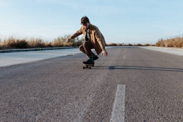 Ein modischer männlicher Skateboarder führt einen bodenberührenden Trick aus, während er sanft über eine Asphaltstraße gleitet - ADSF46449