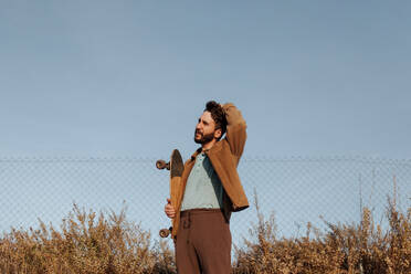 Ein fröhlicher Mann mit Bart und Skateboard steht an einem Zaun und fährt sich lässig mit der Hand durch die Haare - ADSF46455