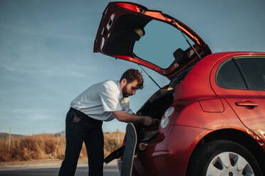 Ein junger Mann bereitet sich auf seine Reise vor, während er neben seinem Auto mit offenem Kofferraum steht, gekleidet in Freizeitkleidung - ADSF46459