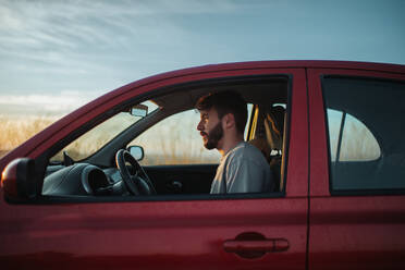 Bearded young male driver in white shirt sitting in car parked on roadside in countryside - ADSF46464