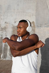 Serious African American athlete in modern headphones warming up before active running training on street - ADSF46473